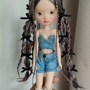 May include: A doll with gray braided hair adorned with black bows. The doll wears a denim crop top and shorts, paired with white socks. The doll has rosy cheeks and a neutral expression.