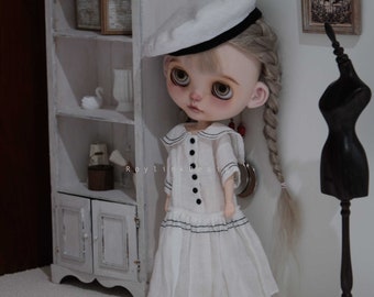 Può includere: Una bambola bianca con trecce bionde che indossa un vestito da marinaio bianco con bordi neri e un cappello da marinaio bianco. La bambola è in piedi davanti a un armadio bianco con ripiani.