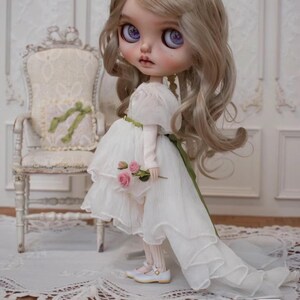 May include: A porcelain doll with long blonde hair wearing a white dress with a green sash and holding a bouquet of pink roses. The doll is standing on a white lace tablecloth.