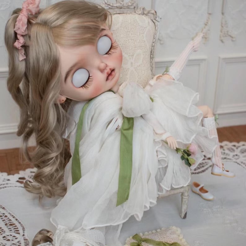 May include: A porcelain doll with blonde hair and a white dress with green ribbon accents sits on a white chair. The doll has large, white eyes and a delicate facial expression.