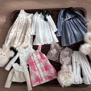 May include: A collection of doll clothes, including dresses, tops, and pants, in various colors and patterns. The dresses feature floral prints, gingham, and lace details. Accessories include a bunny-shaped hat and mittens.
