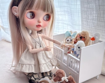 May include: A Blythe doll with long blonde hair and a beige dress, wearing a decorative headband. The doll is surrounded by miniature stuffed animals, including teddy bears and dogs, and a white crib.