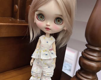 Puede incluir: Una muñeca Blythe con cabello rubio claro y grandes ojos verdes expresivos. La muñeca viste un conjunto blanco con detalles coloridos y ribete de encaje, que incluye una blusa, pantalones cortos y calcetines. Un lazo a juego adorna el cabello de la muñeca.