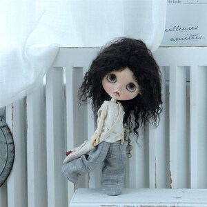 May include: A doll with long, curly black hair wearing a white shirt and gray pants. The doll is holding a brown bag in its hand and is standing on a white shelf.