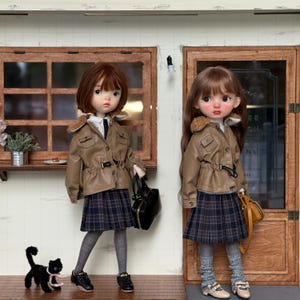 May include: Two dolls dressed in school uniforms stand in front of a wooden facade. They wear brown jackets with faux fur collars, plaid skirts, and sneakers. One doll holds a black purse, and the other a tan purse. A black cat figurine with a pink scarf is in front of one doll.