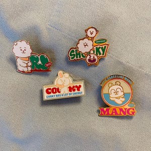 May include: Four colorful enamel pins featuring BT21 characters. One pin shows a white fluffy character with a scarf next to a green shape with the text "BT21". Another pin has a monkey and cloud with the text "Shooky". A third pin has a bunny with the text "Cooky". The last pin has a bear with the text "Mang".