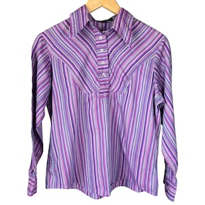 May include: A long-sleeved, button-down shirt with vertical stripes in shades of purple, pink, and white. The shirt features a collared neckline and a button placket. The garment is displayed on a wooden hanger against a white background.