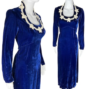 May include: A long, royal blue velvet dress with long sleeves. The dress features a high, rounded neckline trimmed with white floral lace. The dress is displayed on a mannequin.