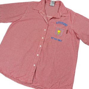 May include: Red and white checkered button-up shirt with short sleeves. The shirt has a collar and a pocket on the left chest with the words "ATTITUDE" and "WE MAY SELF" embroidered in blue. A yellow cartoon character is also embroidered on the pocket.
