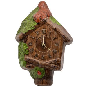 May include: A brown ceramic cuckoo clock shaped like a house, with a squirrel on top and green leaves. The clock face has Roman numerals and gold-colored hands. A ladybug is on a leaf near the base.