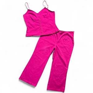 May include: A bright pink two-piece outfit, consisting of a camisole top and matching pants. The top has thin straps and a fitted bodice. The pants are wide-legged. The set is made of a solid-colored fabric.