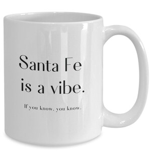 May include: White ceramic coffee mug with black text that reads "Santa Fe is a vibe. If you know, you know."