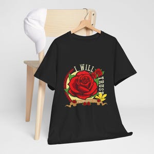 May include: Black t-shirt featuring a large red rose graphic with the words "I WILL TRUST" and "PROVERBS 3:5". The design includes a yellow oval, a banner, and additional red roses. A white hat rests on a wooden chair.