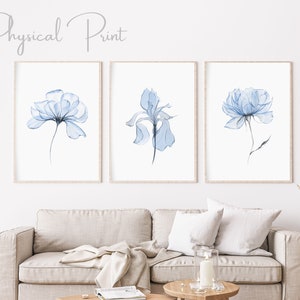 Op de afbeelding: Drie ingelijste kunstdrukken met aquarel illustraties van blauwe bloemen. De prints worden weergegeven boven een beige bank. De tekst "Physical Print" staat bovenaan de afbeelding.