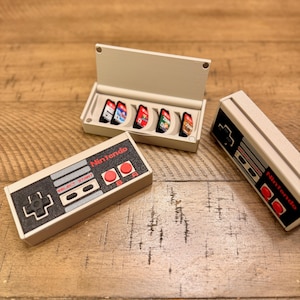 May include: A collection of Nintendo-themed items, including a controller and a case with game cartridges. The controller features a black and red design with the Nintendo logo. The case is open, revealing several colorful game cartridges. The items are on a wooden surface.