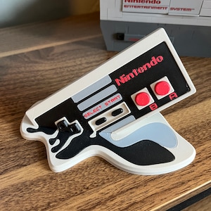 Puede incluir: Un mando de control blanco de Nintendo Entertainment System con botones rojos y un diseño en blanco y negro. El mando tiene forma de pistola y lleva la inscripción "Nintendo".