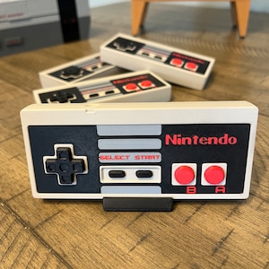 Op de afbeelding: Een witte en zwarte Nintendo-controller met rode knoppen. De controller heeft een directionele pad, select- en startknoppen, en A- en B-knoppen. De controller ligt op een houten oppervlak.