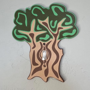 Puede incluir: Una tapa de interruptor de luz con forma de árbol con hojas verdes y un tronco marrón. El interruptor es blanco y está en la posición "encendido".