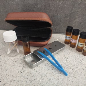 May include: A brown zipped carrying case, a clear plastic jar with two cotton swabs, four small amber glass vials labelled Birch, Anise, Cypress, and Clove, a rectangular metal tin with a sliding lid, and a pair of bright blue plastic tweezers.