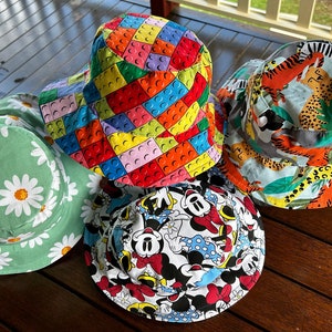May include: Four colourful bucket hats with different patterns. One hat has a green background with white daisies, another has a white background with a multi-coloured Lego pattern, another has a white background with a multi-coloured Mickey Mouse pattern, and the last hat has a light blue background with a multi-coloured animal print.