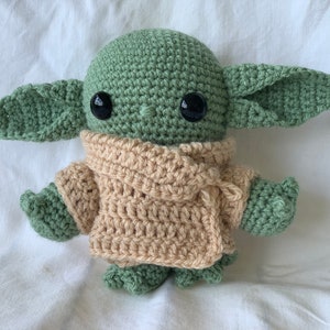 May include: A crocheted green and beige plush toy resembling a popular fictional character. The toy has large black eyes and a beige crocheted scarf.