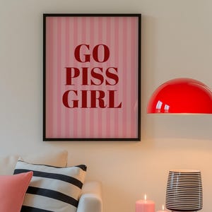 May include: A framed pink and white striped print with the words "GO PISS GIRL" in red lettering hangs above a white sofa with pink and black and white striped pillows. A red lamp and side table with candles and a black and white striped vase are also in the image.