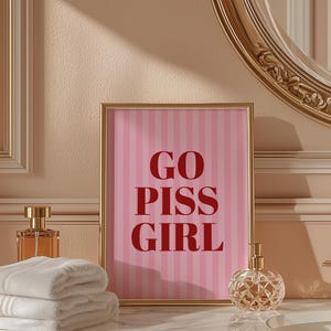 May include: A framed pink and white striped print with the words "GO PISS GIRL" in red lettering. The print is on a marble countertop with a gold-framed mirror, perfume bottles, and folded white towels.