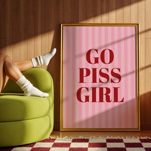 May include: A framed pink and white striped poster with the words "GO PISS GIRL" in red. The poster is in a gold frame and is leaning against a wooden wall. A green sofa, a red and white checkered rug, and a glass lamp are also visible.