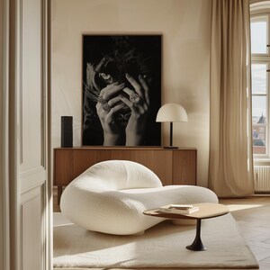 May include: A white, plush, modern armchair with a curved design sits in a room with a wooden cabinet and a black and white print of a panther. A small, round, wooden table with a book on top sits in front of the chair.