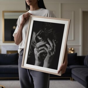 May include: Black and white photograph of a woman's hands holding a black panther's face. The panther's eyes are visible, and the woman is wearing several rings with large gemstones.