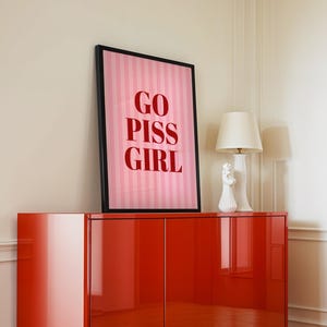 May include: A framed pink and white striped print with the words "GO PISS GIRL" in red lettering. The artwork is displayed on a glossy red cabinet, with a white lamp and decorative figurine.
