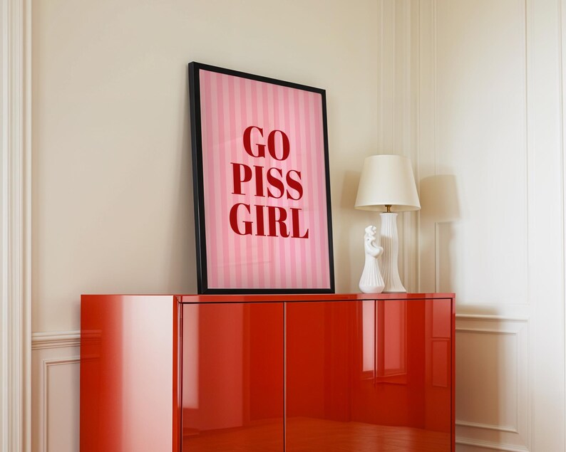 May include: A framed pink and white striped print with the words "GO PISS GIRL" in red lettering. The artwork is displayed on a glossy red cabinet, with a white lamp and decorative figurine.