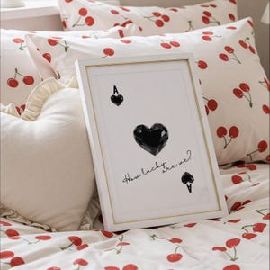 May include: A white framed print with a black heart and the text "How lucky are we?"  The print is on a white bed with a red cherry pattern.