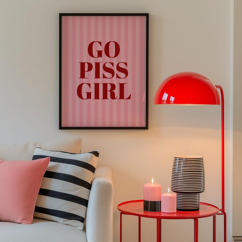 May include: A framed pink and white striped print with the words "GO PISS GIRL" in red lettering hangs above a white sofa with pink and black and white striped pillows. A red lamp and side table with candles and a black and white striped vase are also in the image.