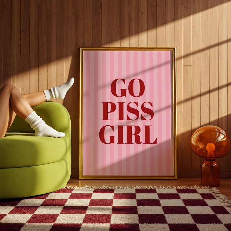 May include: A framed pink and white striped poster with the words "GO PISS GIRL" in red. The poster is in a gold frame and is leaning against a wooden wall. A green sofa, a red and white checkered rug, and a glass lamp are also visible.