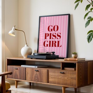 May include: A mid-century modern style wooden media console with a framed pink and white striped poster that reads "GO PISS GIRL". A record player sits on top, with a white lamp and vase to the left.