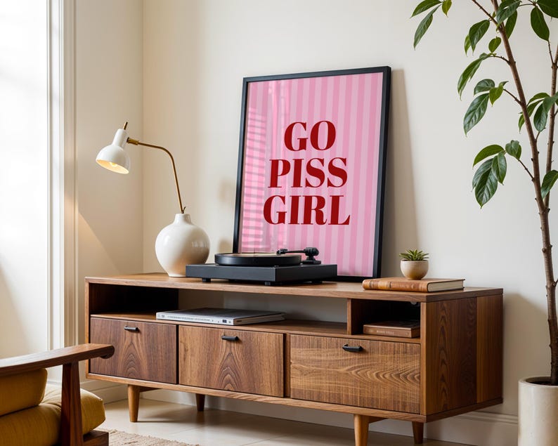 May include: A mid-century modern style wooden media console with a framed pink and white striped poster that reads "GO PISS GIRL". A record player sits on top, with a white lamp and vase to the left.