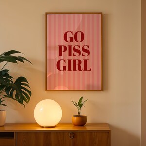 May include: A framed pink and white striped poster with the words "GO PISS GIRL" in red lettering hangs on a beige wall. A round, glowing lamp sits on a wooden cabinet, with a potted plant to the left and a small plant in a terracotta pot to the right.
