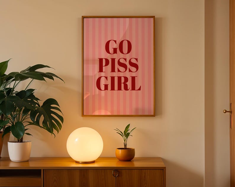 May include: A framed pink and white striped poster with the words "GO PISS GIRL" in red lettering hangs on a beige wall. A round, glowing lamp sits on a wooden cabinet, with a potted plant to the left and a small plant in a terracotta pot to the right.