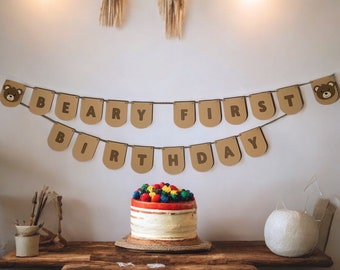 May include: A brown paper banner with the words "Beary First Birthday" hangs above a cake with colorful frosting and berries. The banner has a brown bear face on each end.