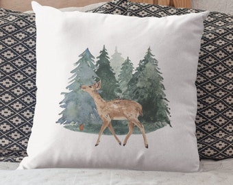 Könnte beinhalten: Ein weißes Kissen mit einer Aquarell-Illustration eines Rehs, das durch einen Wald geht. Das Reh ist braun und die Bäume sind grün.