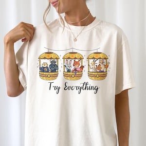 Pode incluir: Camiseta creme com um gr&aacute;fico extravagante de tr&ecirc;s g&ocirc;ndolas, cada uma com animais de desenho animado. As g&ocirc;ndolas est&atilde;o suspensas por uma linha, e o texto "Try Everything" est&aacute; impresso abaixo.