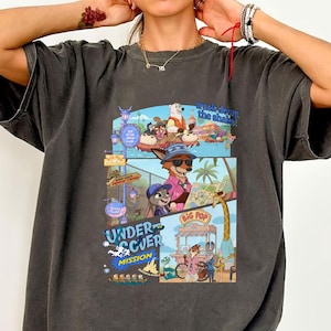 May include: Dark gray t-shirt featuring a colorful cartoon graphic with the words "Undercover Mission" and "Track Down the Shakes". The graphic depicts various cartoon characters in a comic book style.