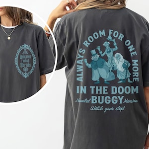 May include: Dark gray t-shirt with a teal graphic on the back featuring three cartoon figures and the text "ALWAYS ROOM FOR ONE MORE IN THE DOOM BUGGY Mansion". The front of the shirt has a teal oval design with the words "Welcome Foolish Mortals".