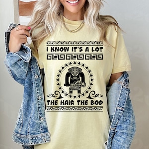 May include: A yellow t-shirt with a black graphic of a cartoon character with the text "I know it's a lot the hair the bod" printed on it.