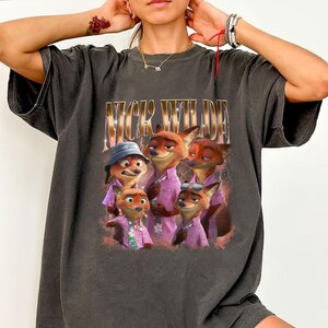 May include: Dark gray t-shirt featuring a graphic of Nick Wilde from Zootopia. The image shows Nick Wilde and other fox characters wearing pink shirts. The text "NICK WILDE" is printed above the image in gold.
