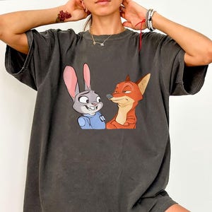 May include: Dark gray t-shirt featuring a cartoon image of Judy Hopps the bunny and Nick Wilde the fox from Zootopia. Judy is wearing a blue uniform, and Nick is orange. The shirt has a vintage, worn look.