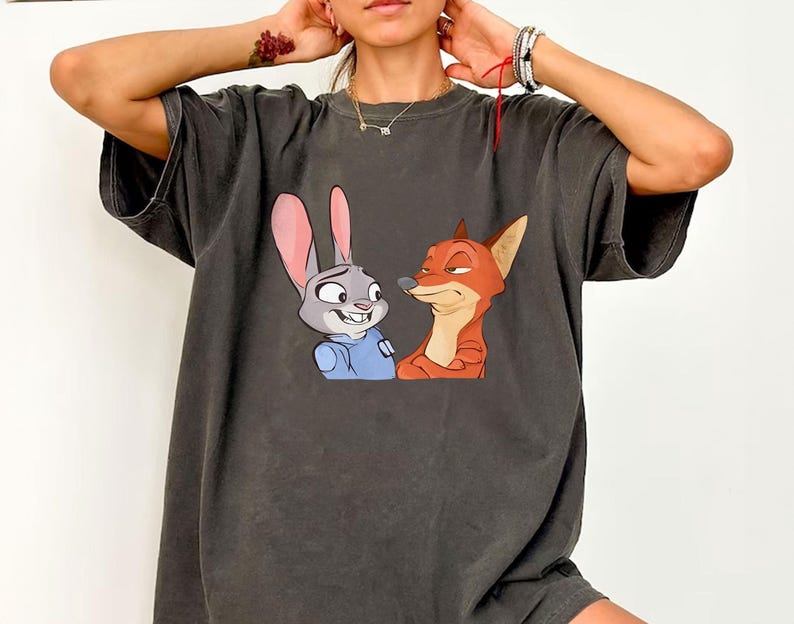May include: Dark gray t-shirt featuring a cartoon image of Judy Hopps the bunny and Nick Wilde the fox from Zootopia. Judy is wearing a blue uniform, and Nick is orange. The shirt has a vintage, worn look.