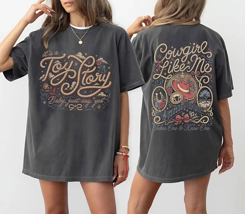 May include: A charcoal gray t-shirt featuring a front graphic with rope-style text reading It's a Toy Story Baby just say yes and a back graphic with the text Cowgirl Like Me Takes One to Know One, a red cowboy hat, horseshoe, and floral motifs.