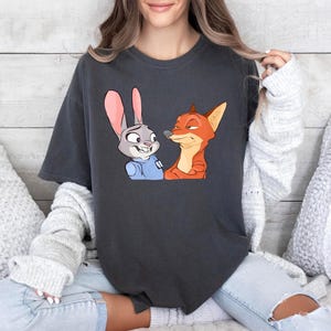 May include: Dark gray t-shirt featuring a cartoon illustration of a rabbit and a fox. The rabbit has gray fur and pink ears, and the fox is orange. The characters are from the animated movie Zootopia.